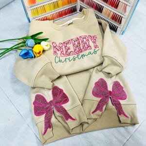 May include: A beige Christmas sweatshirt and matching trousers set. The sweatshirt has the text "MERRY Christmas" in pink and green embroidery. The trousers and sleeves have large pink glitter bow appliques.