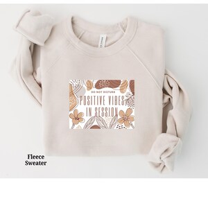 May include: A beige fleece sweatshirt with a graphic print that says "Do Not Disturb, Positive Vibes in Session". The graphic is surrounded by abstract floral shapes in brown, beige, and white.