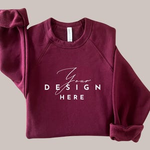 以下が含まれることがあります： 白のクルーネックと長袖のワインレッドのトレーナー。トレーナーには「Your Design Here」という文字が前面に印刷されています。