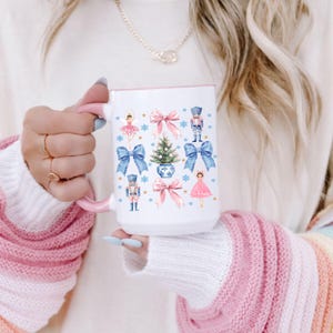 May include: White ceramic mug with a pink handle and rim, decorated with watercolor illustrations of a Christmas tree, bows, nutcrackers, and a ballerina. The mug is held by a person wearing a white sweater.