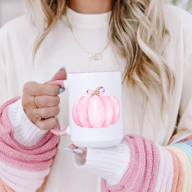 May include: White ceramic mug with a pink interior and handle. The mug features a watercolor illustration of a pink pumpkin with a bow. The mug is held by a person wearing a pink and white sweater.