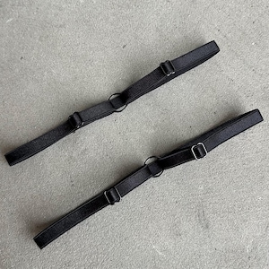 May include: Two black garter belts with adjustable straps and metal hardware. Each belt features a circular ring and two adjustable sliders. The belts are lying on a gray surface.