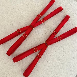 May include: Two sets of red lingerie straps with gold-colored metal hardware. Each set has two straps that cross and connect with adjustable rings. The straps are made of a smooth, satin-like material.