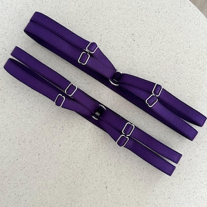 May include: Two sets of purple lingerie straps with silver-colored metal hardware. Each set has two parallel straps connected by a ring and adjustable sliders. The straps are made of a smooth, satin-like material.