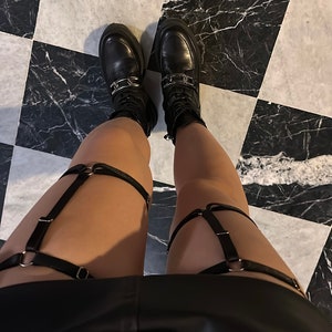 May include: A pair of black leather-look thigh garters featuring adjustable straps, silver-toned metal rings, and sliders. The garters are worn over the legs, paired with black lace-up boots that have a thick sole and a silver-toned metal chain detail.