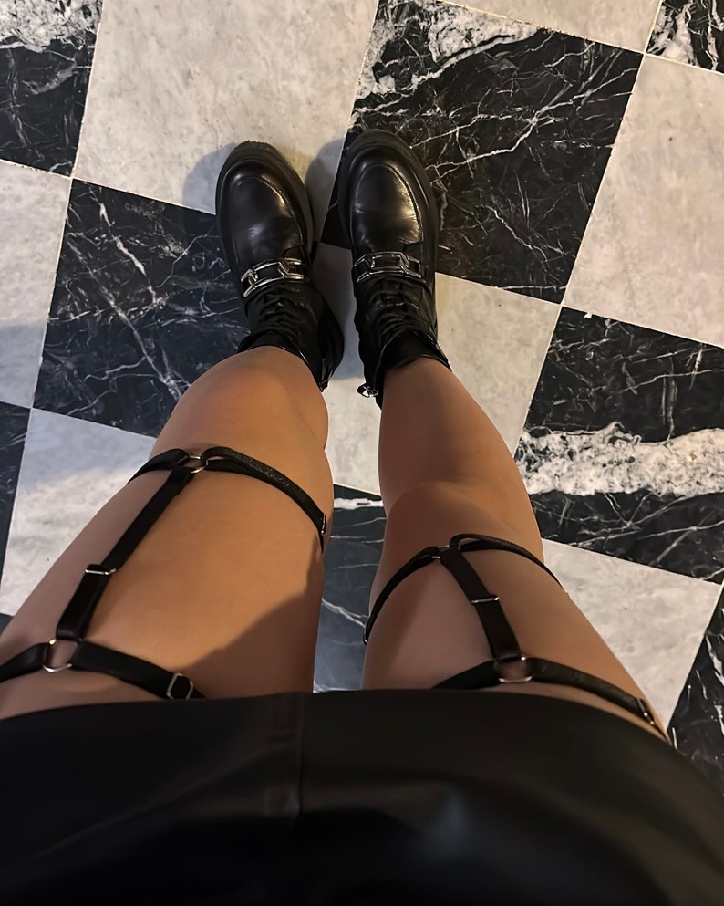 May include: A pair of black leather-look thigh garters featuring adjustable straps, silver-toned metal rings, and sliders. The garters are worn over the legs, paired with black lace-up boots that have a thick sole and a silver-toned metal chain detail.