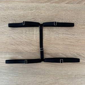 May include: A black elastic leg garter featuring two horizontal bands connected by a vertical center strap. The design includes adjustable silver-toned metal sliders and circular rings that join the elastic straps together in an H-shaped configuration.