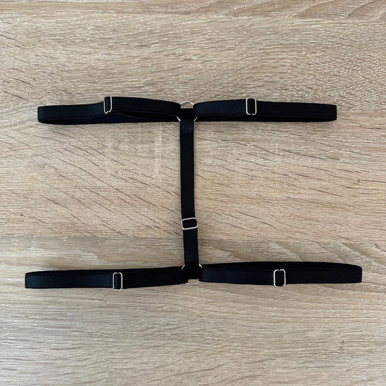 May include: A black elastic leg garter featuring two horizontal bands connected by a vertical center strap. The design includes adjustable silver-toned metal sliders and circular rings that join the elastic straps together in an H-shaped configuration.