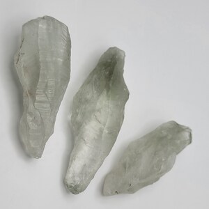 May include: Three light green crystal points with a clear, translucent appearance. The crystals are irregularly shaped and have a rough, natural texture.