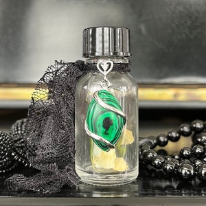 Könnte beinhalten: Eine klare Glasflasche mit schwarzem Deckel und einem silbernen Anhänger, der am Deckel hängt. Der Anhänger ist ein Herz mit einem grünen Malachit-Stein im Inneren. Die Flasche ist mit einer klaren Flüssigkeit gefüllt und am Boden befinden sich kleine weiße Kristalle.