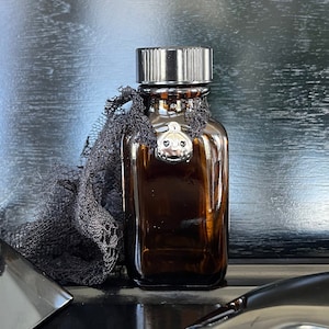 May include: A dark brown glass bottle with a silver lid and a silver charm. The bottle is sitting on a black surface with a black pyramid and a black stone.