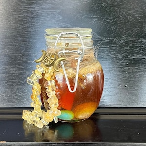 May include: A glass jar filled with a golden liquid and colorful stones, topped with a metal lid and a charm with a crescent moon and pentagram design. A yellow beaded necklace is draped over the jar.