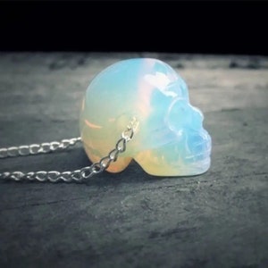 Op de afbeelding: Een witte opaal schedel hanger aan een zilveren ketting. De schedel is gesneden uit een doorschijnende opaal steen die iriserende kleuren vertoont.