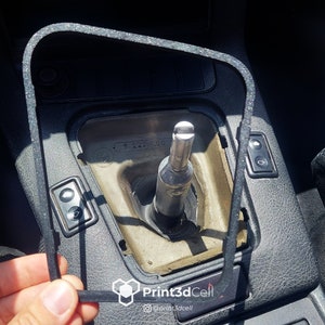 Puede incluir: Primer plano de una junta de goma negra para la palanca de cambios de un coche. La junta tiene forma de rectángulo con esquinas redondeadas. La junta está siendo sostenida por una mano.