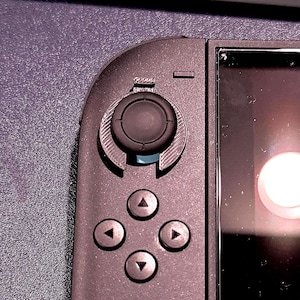 May include: Close-up of a black handheld gaming device. The device features a circular joystick, directional buttons, and a screen. The joystick is surrounded by a curved design. The buttons are in a diamond formation. The screen is reflective.