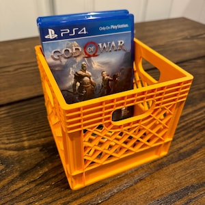 Könnte beinhalten: Ein orangefarbener Plastikkasten mit einem Videospiel-Etui im Inneren. Das Spiel-Etui ist für das PlayStation 4-Spiel "God of War". Das Spiel-Etui hat einen blauen Hintergrund mit dem Spieltitel in Weiß und dem PlayStation-Logo in Weiß.
