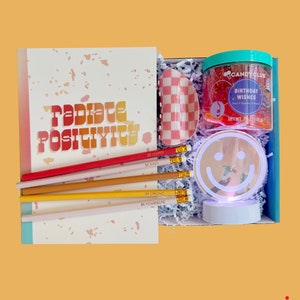 May include: A gift box filled with various items, including a notebook with the text "radiate positivity", a set of pencils with motivational messages, a pink and white checkered hair clip, a clear jar of slime with the text "Candy Club Birthday Wishes", and a white light-up smiley face.