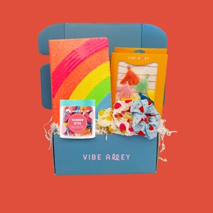 May include: A blue box filled with colorful items. Includes a notebook with a rainbow design, a jar of "Rainbow Bites" candy, a set of tassels, and patterned scrunchies. The box has the text "VIBE ALLEY".