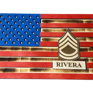 May include: Wooden American flag with blue stars and red stripes, featuring a Sergeant rank insignia and the name "RIVERA". The flag is made of wood with a burnt wood effect.