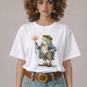 Puede incluir: Camiseta blanca con una ilustración caprichosa de una rana con traje sosteniendo una flor. La rana lleva sombrero, chaqueta y chaleco. La modelo lleva cinturón marrón y falda vaquera.