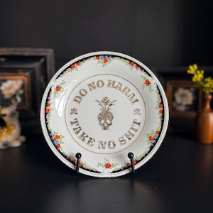 May include: White decorative plate with a gold rim and floral border. The plate features the text "DO NO HARM TAKE NO SHIT" around a gold glitter skull and pineapple design. The plate is displayed on a black metal stand.