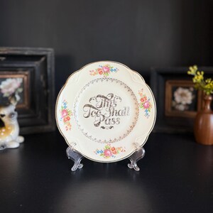 May include: A vintage cream-colored plate with a scalloped edge, adorned with floral patterns in pink, blue, and yellow. The plate features the text "This Too Shall Pass" in a decorative font, with gold accents.