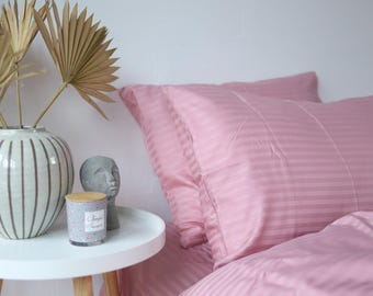 May include: A bedroom setting with a pink striped duvet cover and pillow set. A white side table displays a decorative vase with dried palm leaves, a grey bust, and a scented candle. The bedding is neatly arranged, creating a calm atmosphere.