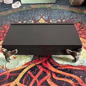 May include: A black wooden box with decorative metal bird feet. The box is rectangular and sits on a colorful puzzle with a tree of life design. The box is likely used for storage or display.