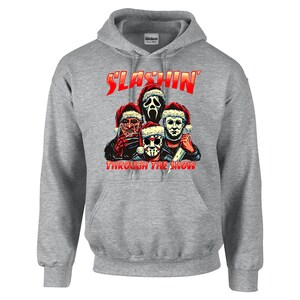 May include: Gray hooded sweatshirt with a red graphic of four horror movie characters wearing Santa hats. The text "SLASHIN' THROUGH THE SNOW" is printed below the characters.
