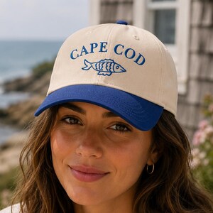 Könnte beinhalten: Beige Baseballkappe mit blauem Schirm und dem Schriftzug "CAPE COD" in Blau, zusammen mit einer Fischgrafik. Die Kappe wird von einer Person getragen. Der Hintergrund zeigt einen Blick auf das Meer und ein verwittertes Gebäude.