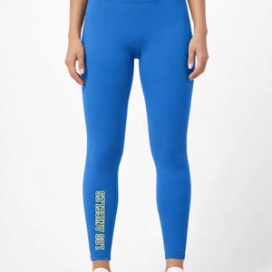 Può includere: Leggings blu brillante con la scritta "LOS ANGELES" stampata verticalmente sulla gamba sinistra in giallo e verde. I leggings sono a vita alta e abbinati a scarpe da ginnastica bianche. L'aspetto generale è atletico e sportivo.