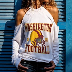 Puede incluir: Sudadera blanca con hombros descubiertos con la inscripción "WASHINGTON FOOTBALL" en burdeos y dorado. Un gráfico de casco de fútbol dorado y burdeos está centrado debajo del texto. La sudadera tiene mangas largas y cuello alto.