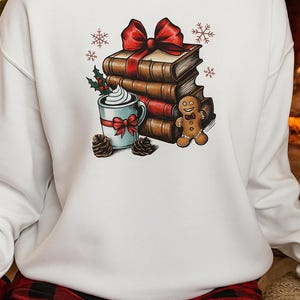 Pode incluir: Camisola branca com um gráfico temático de Natal. O gráfico apresenta uma pilha de livros amarrados com um laço vermelho, uma caneca de chantilly, um homem de gengibre, azevinho, pinhas e flocos de neve. A pessoa está vestindo calças de pijama xadrez vermelho e preto.