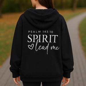 Puede incluir: Sudadera con capucha negra con el texto "SPIRIT lead me" en escritura blanca y un símbolo de corazón. El texto "PSALM 143:10" está encima del texto principal. La persona lleva leggings negros. El fondo está borroso.