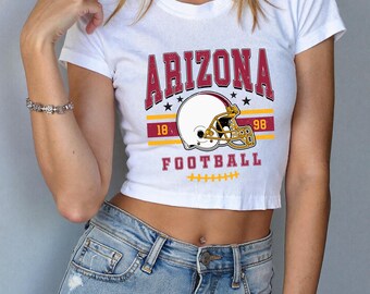 Può includere: Top corto bianco con la scritta "ARIZONA" a blocchi color marrone, una grafica di un casco da football e la scritta "FOOTBALL" in marrone. La maglietta ha anche i numeri "18" e "98" in giallo.