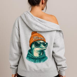 May include: A light gray hoodie featuring a graphic of a dolphin wearing sunglasses and an orange beanie with the word "Miami". The dolphin is wearing a teal hoodie. The hoodie is a casual style.
