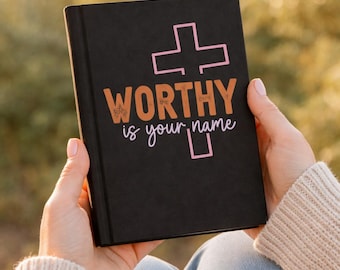 May include: A black journal with the words "WORTHY is your name" in orange and white script, with a pink cross graphic. The journal is held in front of a blurred, natural background.