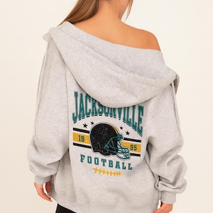 Może przedstawiać: Jasnoszara bluza z kapturem zapinana na zamek z grafiką Jacksonville Football na plecach. Grafika zawiera hełm do futbolu amerykańskiego, napis "JACKSONVILLE" oraz lata "19" i "95".