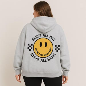 Puede incluir: Sudadera con capucha gris claro con un gráfico de carita sonriente amarilla y el texto "SLEEP ALL DAY NURSE ALL NIGHT". Cuadrados a cuadros en blanco y negro a ambos lados de la carita sonriente.