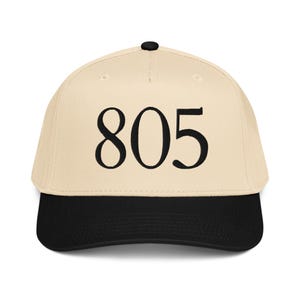 Puede incluir: Gorra de béisbol beige con visera negra y botón negro. El número "805" está bordado en negro en la parte delantera. Este accesorio es elegante.