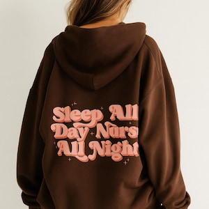 Puede incluir: Sudadera con capucha marrón con el texto "Sleep All Day Nurs All Night" en una fuente retro. La sudadera tiene capucha y un ajuste relajado. El texto es rosa y naranja.