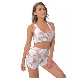 May include: White sports bra and shorts set with a pink ballet-themed pattern. The bra has a halter neckline, and the shorts are ruched. The pattern includes ballet shoes, bows, and ribbons.