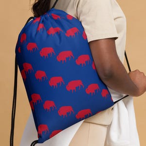 Puede incluir: Una mochila azul con cordón y un patrón repetido de siluetas de búfalos rojos. La bolsa tiene cordones negros. El fondo es de color beige liso.