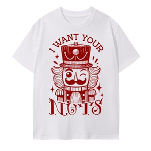 May include: White t-shirt featuring a red graphic of a winking nutcracker with a hat and the text "I WANT YOUR NUTS". The design includes stars and decorative elements, creating a festive and playful look.