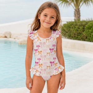 Può includere: Un costume da bagno intero con base bianca e un motivo ripetuto di fiocchi rosa e dorati. Il capo presenta maniche ad aletta con volant e un orlo arricciato lungo le aperture delle gambe, aggiungendo un dettaglio decorativo al design.