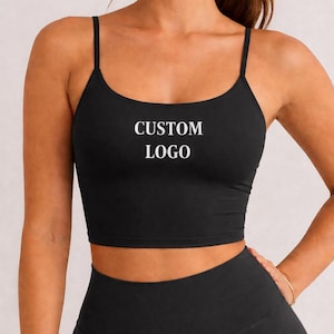 May include: Black crop top with thin straps and the text "CUSTOM LOGO" in white. The top is paired with matching black leggings. The outfit is designed for athletic activities.