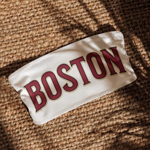 Puede incluir: Top de tubo blanco con la palabra "BOSTON" en letras de bloque granate. La parte superior está hecha de una tela suave y se coloca sobre un fondo texturizado y tejido.