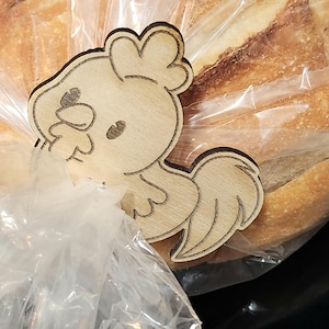 Puede incluir: Un clip para pan con forma de gallo de madera. El clip está hecho de madera de color claro y presenta un diseño de gallo de dibujos animados. El clip sujeta una bolsa de plástico transparente que contiene un pan.