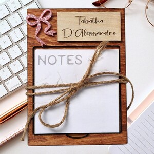 Op de afbeelding: Een houten notitieblokhouder met een roze strik en de naam "Tabatha D'Alessandro" gegraveerd op de bovenkant. Het notitieblok heeft een wit papierblok met het woord "NOTES" erop gedrukt en is vastgebonden met touw.