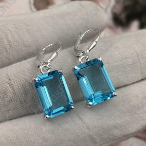 Puede incluir: Un par de pendientes de plata con piedras rectangulares de color azul claro. Los pendientes tienen un cierre de gancho.